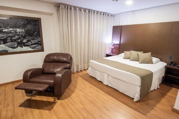 Una habitación de hotel con cama, sillón reclinable, un cuadro en la pared y cortina.