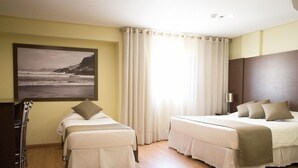 Habitación de hotel con dos camas, un ventanal con cortinas, un cuadro de una playa y un escritorio con sillas.