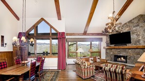 A cozy living room with a stone fireplace, a large TV mounted on the wall, and a dining area with a wooden table and chairs.