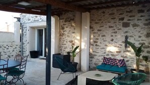 En overdækket terrasse med stenvæg, spisebord med stole, sofa med puder og to krukker med planter.