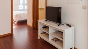 Habitación de hotel con televisor sobre un soporte blanco, cama con cubierta blanca y vista a un balcón a través de una puerta abierta.
