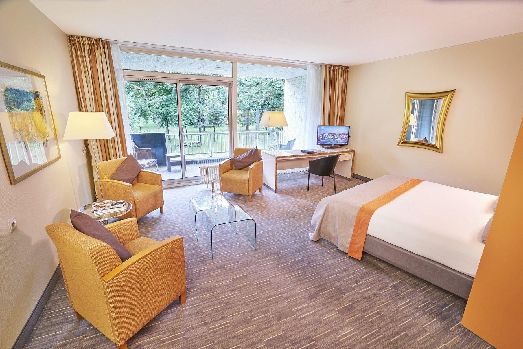 A hotel room with a bed, a desk, two chairs, a television, and a balcony with a view of trees.