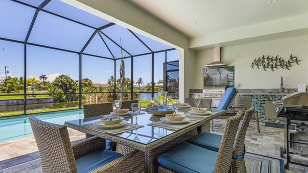 Un área de comedor al aire libre con sillas de mimbre y una mesa de vidrio, con vista a una piscina y un jardín exuberante.