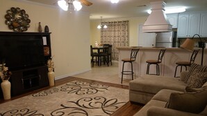 Ein Wohnzimmer mit Sofa, Fernseher und Dekoration. Es gibt eine Küchenzeile mit Bar und Stühlen.