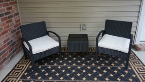 Eine Veranda mit zwei Korbstühlen, einem kleinen Tisch, einem gemusterten Teppich und einer Backsteinmauer.