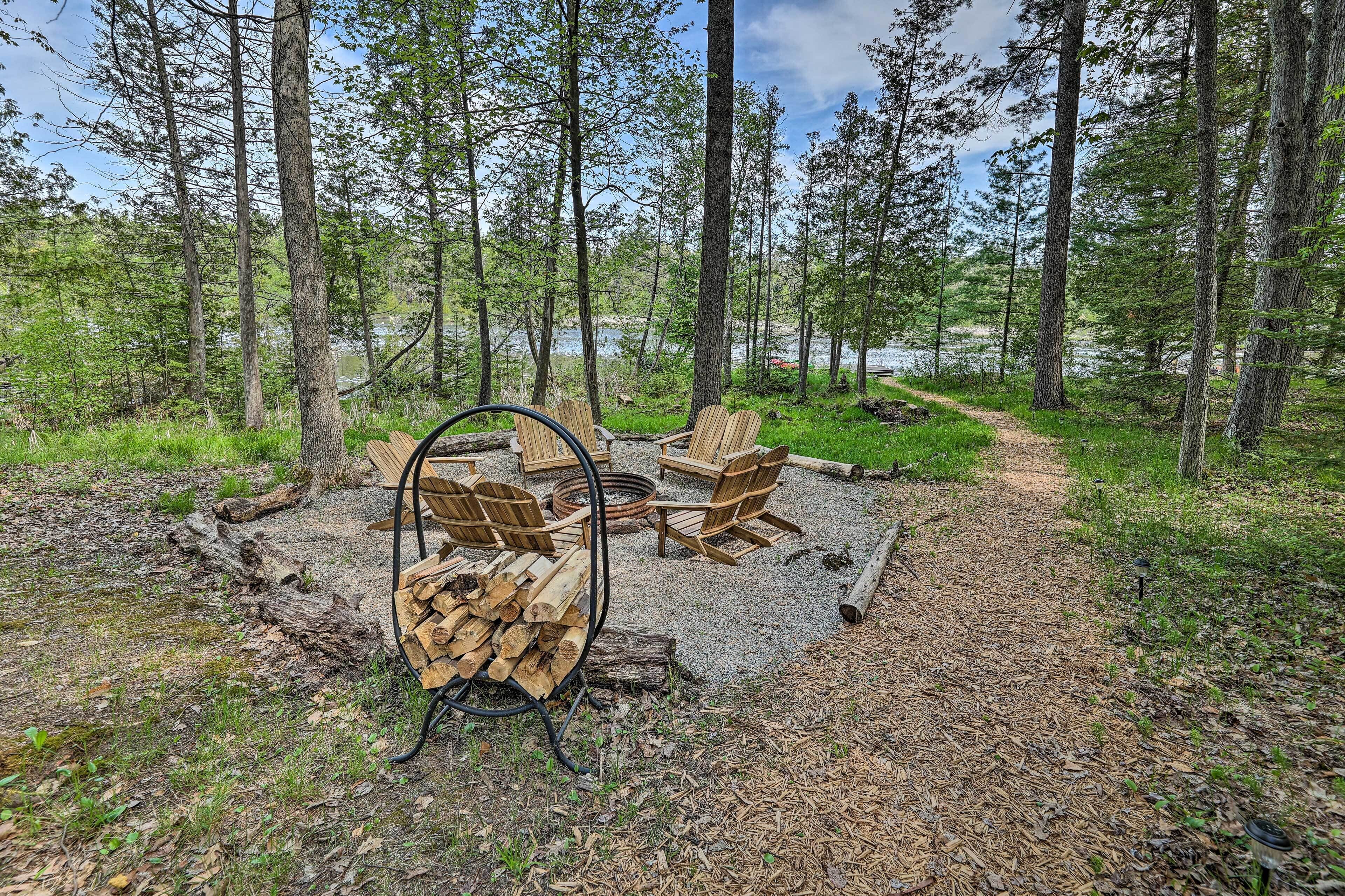 A forested campsite with a fire pit, chairs, and a stack of firewood.
