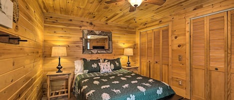 A wooden cabin bedroom with a bed, two bedside lamps, a ceiling fan, a mirror, and wooden closets.