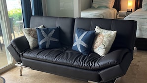 Ein schwarzes Ledersofa mit gemusterten Kissen, ein gefliester Boden und ein Bett mit einem Nachttisch im Hintergrund.