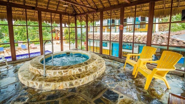 Una piscina interior con paredes de piedra y techo de paja, rodeada de grandes ventanales y sillas amarillas.