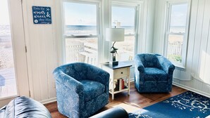 Ein Zimmer mit zwei blauen, gemusterten Sesseln, einem kleinen weißen Beistelltisch mit Lampe und Blick aufs Meer durch große Fenster.