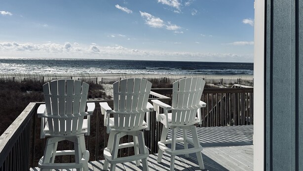 Eine Holzterrasse mit zwei weißen Adirondack-Stühlen, die auf einen Strand ausgerichtet sind.