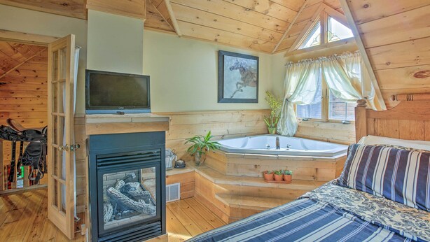 A cozy bedroom with a fireplace, a large window, a bathtub, and a television. - Resort Cabin w/ Fire Pit: Golf, Hike and Play! (Rothbury)