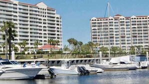 A marina with several boats docked, a large hotel building with multiple balconies, and a clear blue sky.