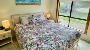 Un dormitorio con cama, una mesita de noche, lámpara, ventilador de techo, ventana y un cuadro con una escena de playa.