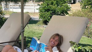 Una persona reclinada en una silla de descanso con un libro, al aire libre con un jardín y una cerca al fondo.
