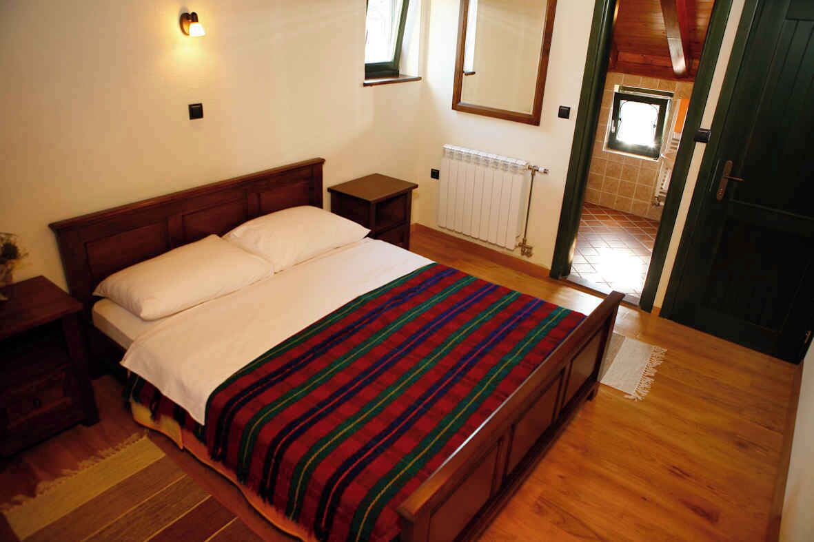 Une chambre à coucher avec un lit en bois, une table de chevet, une commode, un radiateur et une porte donnant sur une autre pièce.