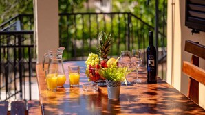 Una mesa de madera con una jarra de jugo de naranja, dos vasos, una botella de vino y un cuenco de fruta en un balcón.