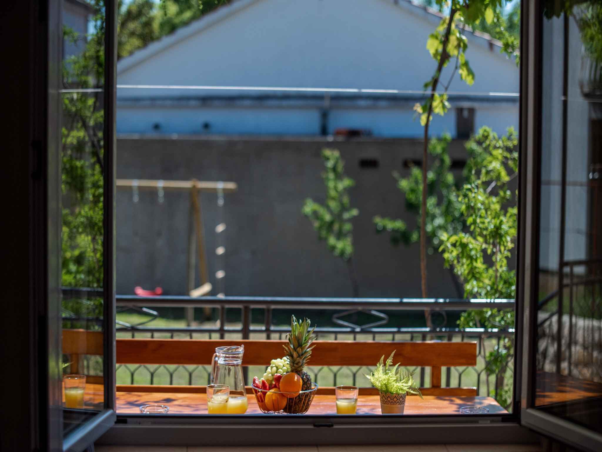 A table with a fruit bowl, pitcher, and glasses on a balcony with a view of a building and trees.
