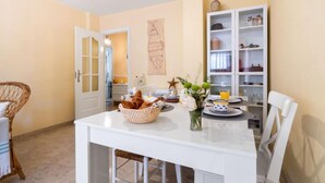 A dining area with a white table set for two, a basket of bread, a vase with flowers, and a shelf with kitchen items. - Rincon Dorado by Interhome (Torremolinos)