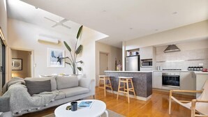 A modern living room with a sofa, coffee table, and a kitchen area with a microwave, oven, and bar stools. - Jacaranda Lane | Sleek & Modern | Central (Margaret River)