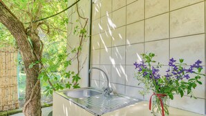 A kitchen with a stainless steel sink, a red-ribboned vase with purple flowers, and a tree trunk leaning against the wall. -  Kipi Village Zakynthos (Ζάκυνθος)