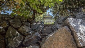 Eine Steintreppe führt zu einem Gazebo mit Ziegeldach, umgeben von großen Steinen und Bäumen.