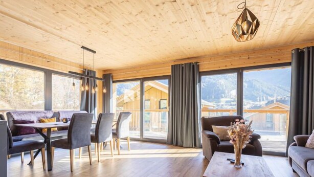 A modern interior with a wooden ceiling, a dining area with a table and chairs, a sofa, and a large window offering a mountain view.