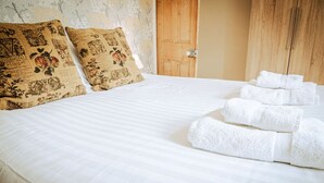 A neatly made bed with white linens, two decorative pillows with floral patterns, and a stack of white towels on the bed. - Painter`s Cottage, a charming cottage in historic Town of Matlock Bath (Derbyshire)