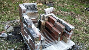 A brick structure with a metal grate, surrounded by grass and scattered bricks. - Loghouse kizuki (Tsuru)
