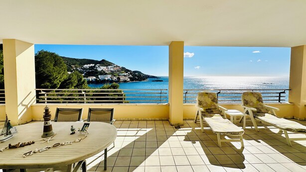 Un balcon avec vue sur la mer et une colline. On y trouve des chaises et une table, ainsi qu'une maquette de bateau et un cadre photo.