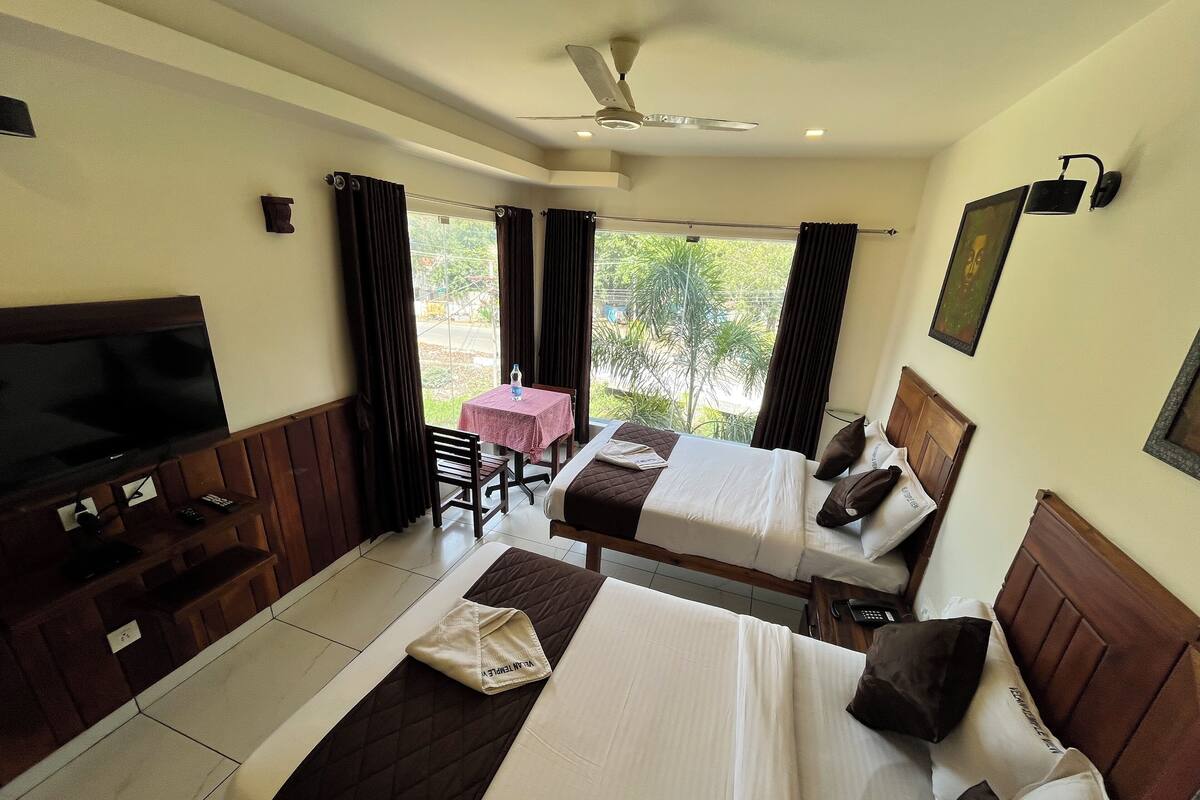 A hotel room with two beds, a TV, a table with a pink tablecloth, and a view of greenery outside.