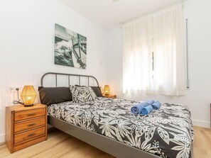 Une chambre à coucher avec un lit, deux lampes de chevet, une table de nuit en bois, un tableau accroché au mur et une fenêtre ornée de rideaux légers.
