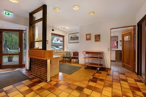 Un espace d’accueil comprenant un comptoir d’enregistrement, une cheminée, une fenêtre avec vue, une porte donnant sur la cuisine et un sol carrelé.