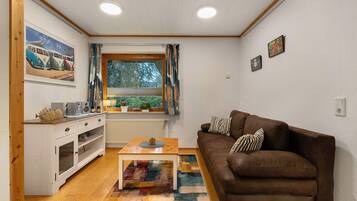 Un soggiorno con un divano marrone, un tavolino in legno, un armadietto bianco, un tappeto colorato, una finestra con tende e un quadro raffigurante auto d'epoca.