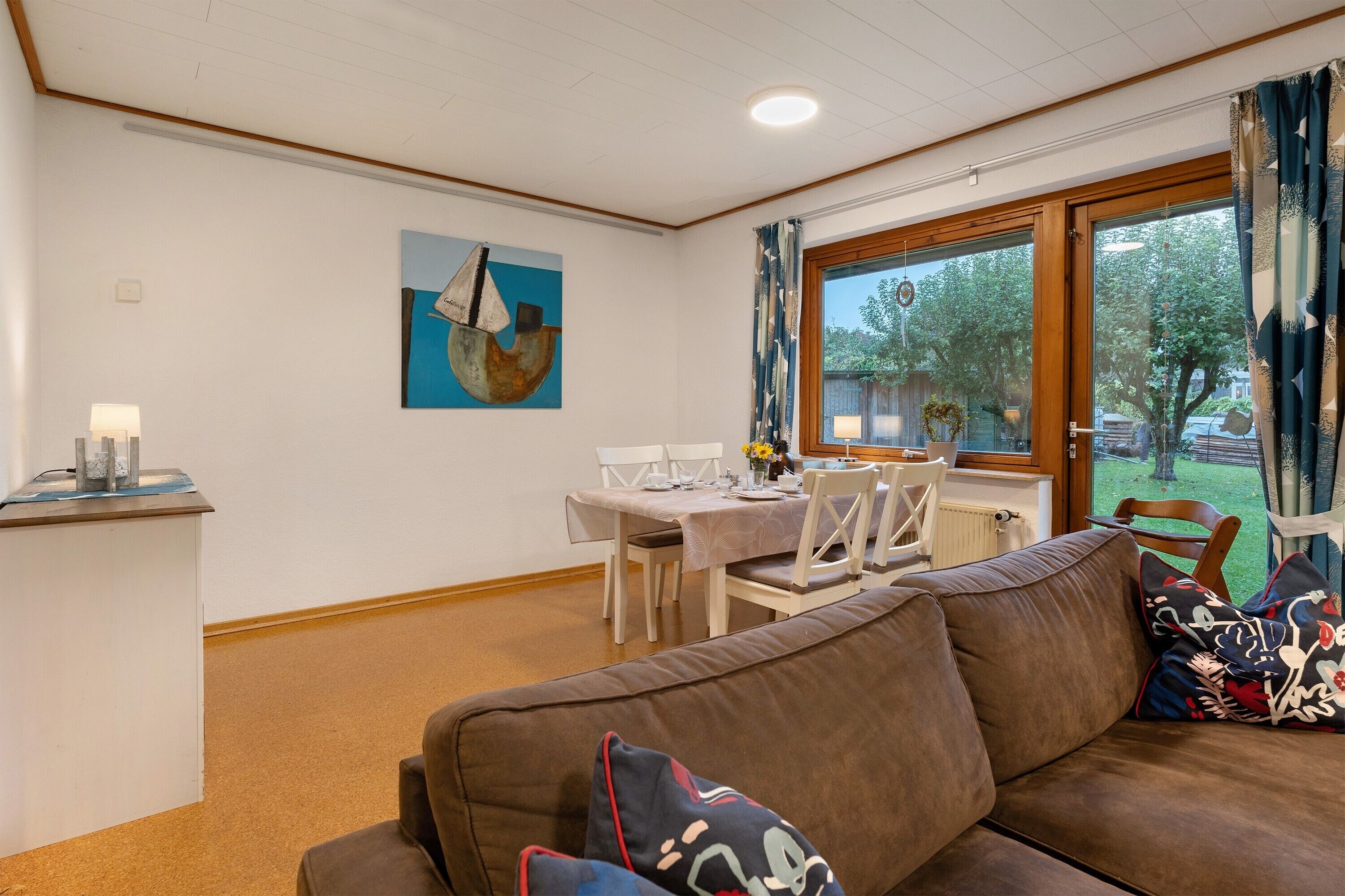 Ein Wohnzimmer mit braunem Sofa, einem Esstisch mit Stühlen, einem Bild an der Wand und einem Fenster mit Vorhängen.