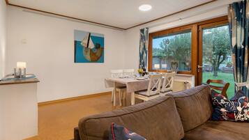Ein Wohnzimmer mit braunem Sofa, einem Esstisch mit Stühlen, einem Bild an der Wand und einem Fenster mit Vorhängen.