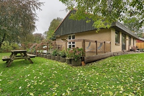 Un bâtiment de style rustique, avec une façade en briques, une terrasse en bois et une table de pique-nique, le tout situé dans un cadre verdoyant.
