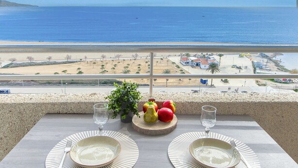 Ein Balkontisch mit zwei Tellern und zwei Gläsern, mit Blick auf einen Strand und einen Parkplatz.