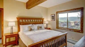 A bedroom with a wooden bed frame, two bedside tables, a window with a view, and a ceiling with exposed beams. - Free EV Charger in 2 Car Garage-3 enSuites-EZ Walk to Resort/Free Town Bus-W/D in Unit-Dogs OK (Steamboat Springs)