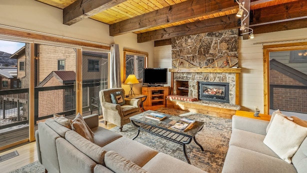 A cozy living room with a stone fireplace, wooden ceiling beams, and a large window overlooking a snowy landscape. - Free EV Charger in 2 Car Garage-3 enSuites-EZ Walk to Resort/Free Town Bus-W/D in Unit-Dogs OK (Steamboat Springs)