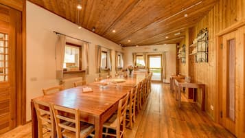 Una larga mesa de comedor de madera con sillas, una zona de cocina con un lavamanos y armarios, y vistas al exterior a través de una ventana.