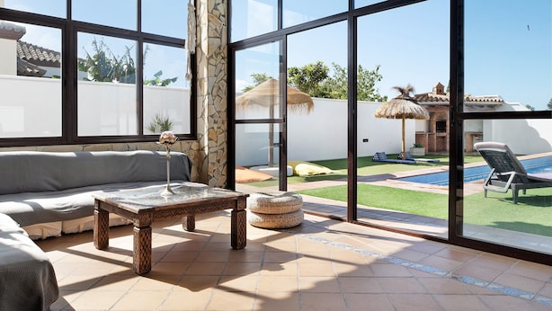 A sunlit room with a large window, a sofa, a coffee table, and a view of a swimming pool and garden. - Chalet "Casa Juan Arias" Close to the Sea with Pool, A/C, Wi-Fi, Garden & Terrace (Conil de la Frontera)