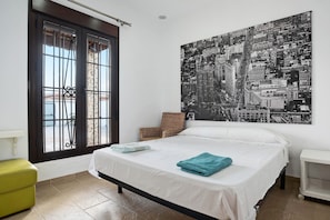 Un dormitorio con una cama, una ventana con barrotes, una silla de mimbre y un cartel con un paisaje urbano en la pared.