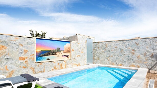 A swimming pool with a stone wall and a mural, lounge chairs, and a clear sky.