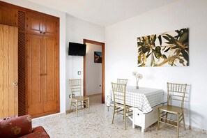 Quarto com guarda-roupa de madeira, televisão na parede, mesa de jantar com cadeiras e um quadro emoldurado.