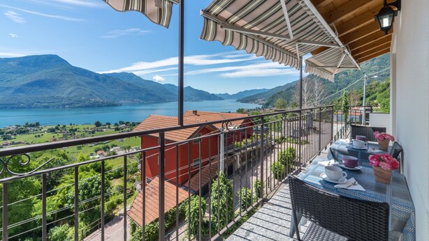 A balcony with a view of a lake and mountains, a table with cups and a vase of flowers, and a red building in the background.