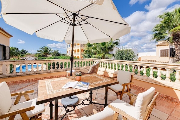 A terrace with a table, chairs, and an umbrella, overlooking a swimming pool and greenery.