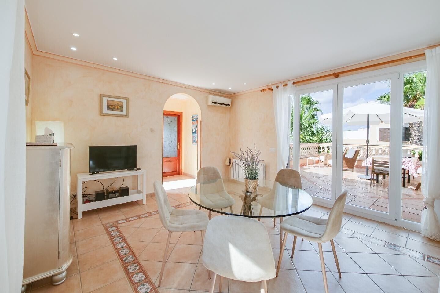 A living room with a TV, a glass table, and chairs, leading to a patio with outdoor furniture.