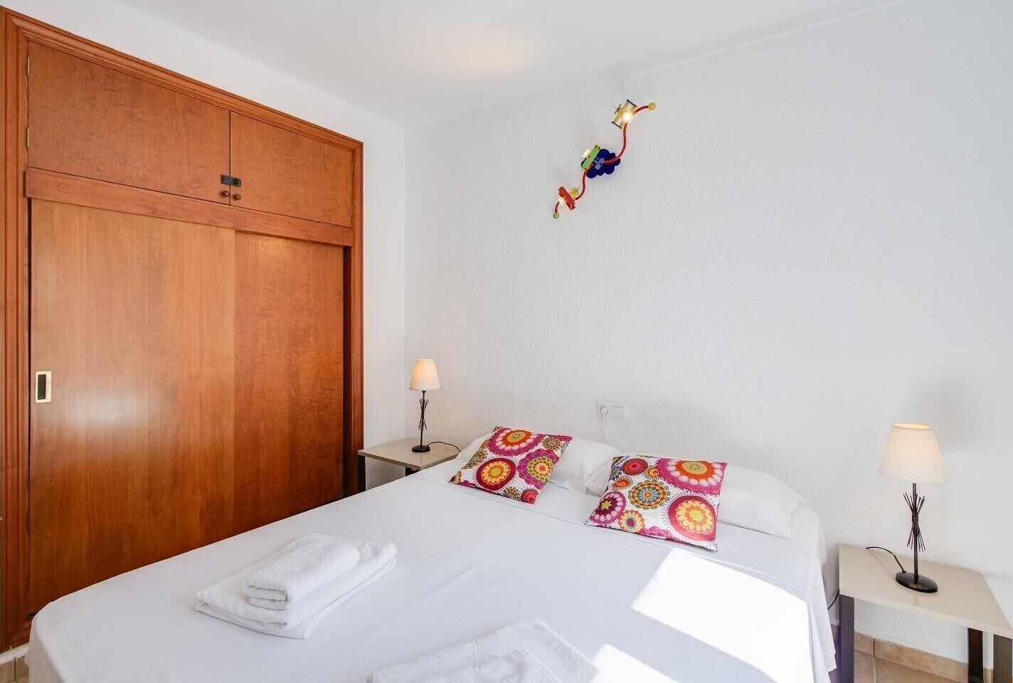A hotel room with a wooden wardrobe, a bed with white linens, colorful pillows, two bedside tables with lamps, and a decorative string of lights.