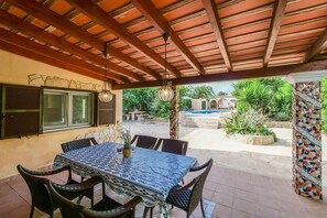 An outdoor patio with a table and chairs, a pool in the background, and a covered area with a wooden ceiling.